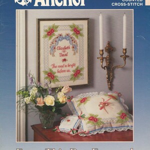 Puede incluir: Una portada para un kit de punto de cruz contado de Anchor. La imagen presenta una pieza de punto de cruz enmarcada con los nombres "Elizabeth" y "David" y el texto "The road is bright before us." También se muestran cojines decorativos y un arreglo floral.