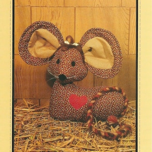 May include: A sewing pattern for a Calico Mouse Doorstop by Honey Bee Patterns, Pattern #D3, 1982 Sharon Wright. The image shows a brown floral fabric mouse with large ears, a red heart patch, and a braided tail, sitting on straw.