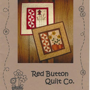 Op de afbeelding: Twee kleine, vierkante quilts met seizoensgebonden thema's worden getoond. De bovenste quilt heeft een rood-wit geometrisch patroon en een vogelhuisje. De onderste quilt heeft pompoenen en bloemen. De tekst "Seasons Two" en "Red Button Quilt Co." zijn zichtbaar.