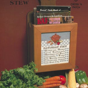 May include: A wooden wall-mounted recipe holder with a framed cross-stitch design that reads "Saturday Stew." The holder contains cookbooks and is decorated with a heart-shaped cutout. Fresh vegetables, including carrots, corn, and mushrooms, are arranged in front.