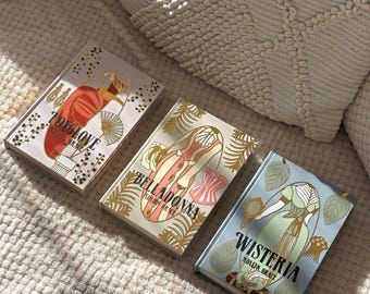 May include: Three hardcover books with decorative covers. The book covers feature illustrations of women in vintage clothing, with titles including "Foxglove", "Belladonna", and "Wisteria". The books are arranged on a textured, off-white surface.