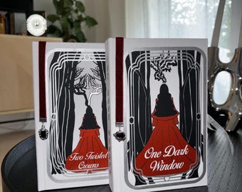 May include: Two hardcover books with matching designs. The covers are white with black and silver art deco style illustrations. The titles are "Two Twisted Crowns" and "One Dark Window". Each book has a red ribbon bookmark.
