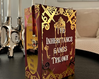 May include: A hardcover book trilogy titled "The Inheritance Games" with a burgundy cover and gold foil accents. The cover features gold illustrations of keys, swords, and a crown. A red ribbon bookmark with a skull charm is visible.