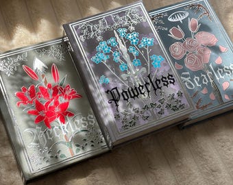 May include: Three hardcover books with decorative covers. The first book is green with red lilies and the text Reckless. The second is purple with blue flowers, a sword, and the text Powerless. The third is blue with pink roses and the text Fearless.