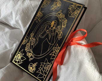 May include: A black book with gold floral and figure illustrations, tied with a bright orange ribbon. The cover features a gold-lined oval with two figures and roses. The book is on a white surface.