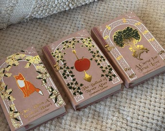 May include: Three hardcover books with pink covers and gold foil accents. The books feature illustrations of a fox, an apple, and a tree. Titles include "Once Upon a Broken Heart", "The Ballad of Never After", and "A Curse for True Love".