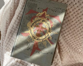May include: A hardcover book titled "The Lord of the Rings" with a gold and red design on the cover. The cover is a muted green with a map-like background. The book is resting on a soft, textured, off-white surface.