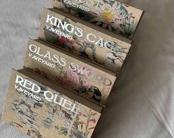 May include: Four decorative boxes with floral and botanical designs. Each box features a different title in silver lettering: "WAR STORY", "KING'S CAGE", "GLASS SWORD", and "RED QUEEN". The boxes are arranged diagonally, showcasing their unique artwork and text.