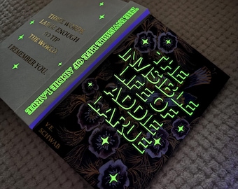 May include: Two hardcover books, "The Invisible Life of Addie LaRue" by V.E. Schwab, with glowing green text and accents. One book is open, revealing the title and author. The other book's cover has the title and floral designs.