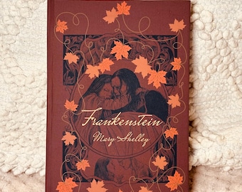 May include: A hardcover book titled "Frankenstein" by Mary Shelley. The cover is a deep reddish-brown with an illustration of two figures embracing within an ornate frame. Orange maple leaves and vines decorate the cover.