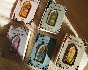 May include: Five decorative books with colorful stained-glass window designs. The book titles include "A Court of Thorns and Roses," "A Court of Mist and Fury," "A Court of Wings and Ruin," "A Court of Frost and Starlight," and "A Court of Silver Flames."