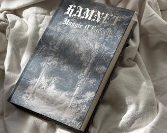 May include: A hardcover book titled "Hamnet" by Maggie O'Farrell. The book's cover features a grayscale illustration of a forest scene. The title is in a gothic font, with the author's name below. The book rests on a white, textured fabric.