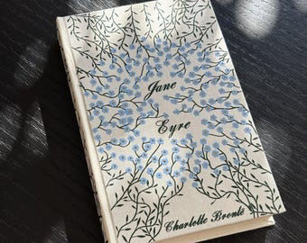 May include: A hardcover book titled "Jane Eyre" by Charlotte Brontë. The book cover is off-white with a floral design of blue flowers and green vines. The title and author's name are in a script font.