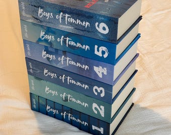 May include: A stack of seven books titled "Boys of Tommen" in various shades of blue and green. The top book has the title "REASING 10" in red. The books are stacked on a white surface.