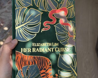 May include: A hardcover book titled "Her Radiant Curse" by Elizabeth Lim. The cover is dark green with gold leaf accents, featuring a red snake, tiger, and tropical leaves. The book is held by a person.