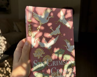 May include: A pink book titled "Six Crimson Cranes" with gold and white crane illustrations. The book has a pink ribbon bookmark with a decorative charm. The cover design features cranes, lotus flowers, and leaves.