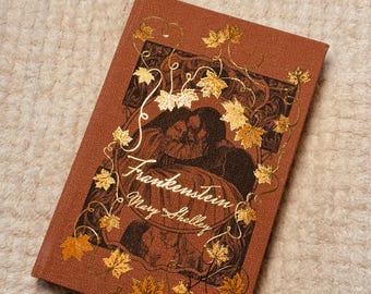 May include: A hardcover book titled "Frankenstein" by Mary Shelley. The book has a brown cover with gold foil accents, including the title and decorative leaves. The cover features a black and brown illustration.