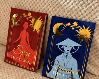 May include: Two hardcover books with gold foil accents. One is red with a woman silhouette and the title "Spin The Dawn". The other is blue with a figure in a hat and the title "Unravel The Dusk". Both have sun and moon designs.