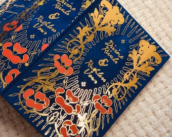 May include: A navy blue book with gold floral designs and the text "Divine Rivals & Ruthless Vows" in gold lettering. The cover features red poppy illustrations. The book is partially open, revealing the inside pages.