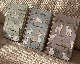 May include: Three hardcover books with pastel covers featuring illustrations and gold lettering. The books are titled "Pride & Prejudice", "Persuasion", and "Sense & Sensibility" by Jane Austen. The covers have illustrations of swans, rabbits, and sheep.