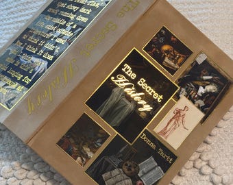 May include: A hardcover book titled "The Secret History" by Donna Tartt. The book is wrapped in a tan cover with gold lettering and decorative images. The cover features a quote and several small framed images.