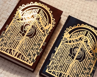 May include: Two hardback books with gold gate and rose designs. One book is dark brown, the other is navy blue. The gold design includes the word "PHANTASMA" on the brown book and "ENCHANTRA" on the blue book.