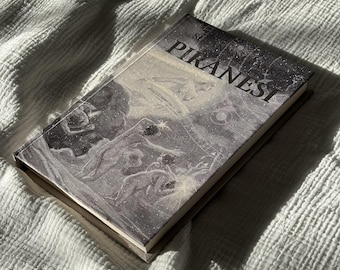 May include: A hardcover book titled "Piranesi" with a grayscale illustration on the cover. The artwork depicts figures in a celestial or dreamlike setting. The book is resting on a white, textured surface.