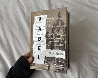 May include: A hardcover book titled "Babel" with a vintage aesthetic. The cover features a sepia-toned illustration of a domed building and handwritten script. The book is held against a white background.