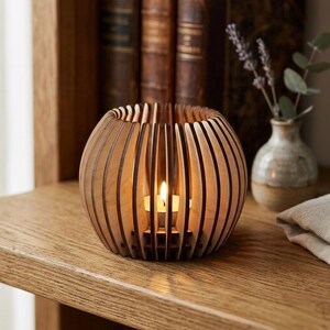 Könnte beinhalten: Ein kugelförmiger Kerzenhalter aus Holz mit vertikalen Lamellen. Im Inneren brennt eine kleine Kerze, die ein warmes Licht ausstrahlt. Der Kerzenhalter steht auf einem Holzregal, im Hintergrund Bücher und eine Vase mit Blumen.