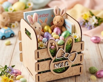 Könnte beinhalten: Ein hölzerner Osterkorb mit Hasenmotiv, gefüllt mit Plüschhasen, Schokoladeneiern und einem Buch mit dem Wort "Easter". Der Korb trägt den Namen "Nele" und ist mit Blumen und Süßigkeiten verziert.