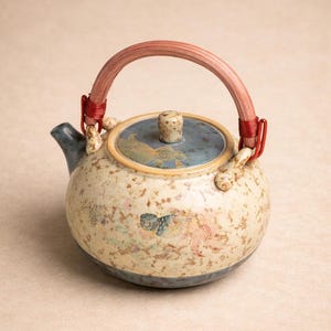Könnte beinhalten: Eine Keramik-Teekanne mit gesprenkeltem cremefarbenem Körper, blauem Deckel und einem gebogenen Holzgriff, der mit rotem Draht umwickelt ist. Die Teekanne hat bemalte Blumen- und Vogelmotive. Ausguss und Boden sind blau.