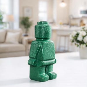 May include: A green decorative figurine in the shape of a classic toy figure. The figurine has a speckled texture and is standing upright. The figure has a head, torso, arms, and legs. The figurine is on a white surface.
