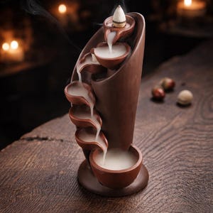 Könnte beinhalten: Ein brauner Keramik-Räucherwasserfall mit kaskadenförmigen Schalen. Oben befindet sich ein angezündeter Räucherkegel, der einen Strom weißen Rauchs freisetzt, der die Schalen hinunterfließt. Der Wasserfall steht auf einer Holzoberfläche.