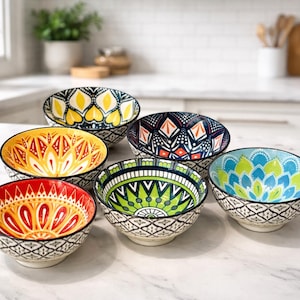 May include: A collection of six ceramic bowls with vibrant, patterned designs. The bowls display floral and geometric motifs in shades of yellow, orange, red, green, blue, and navy. Each bowl has a black and white patterned rim. Ideal for serving or display.
