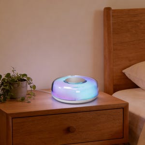 Op de afbeelding: Een witte en zilveren bedlamp met een donutvorm, die een zachte gradiënt van paars, blauw en groen licht uitstraalt. De lamp staat op een houten nachtkastje.