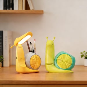 Puede incluir: Dos organizadores de escritorio con forma de caracol. Uno es una lámpara de escritorio amarilla con una concha gris y un portalápices. El otro es un portalápices verde y amarillo con un diseño en espiral en la concha. Ambos están sobre un escritorio de madera.
