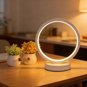 Puede incluir: Una moderna lámpara de anillo LED blanca con un diseño circular, colocada sobre una base redonda. La lámpara está sobre una mesa de madera, junto a dos pequeñas macetas de cerámica blanca con plantas decorativas. La lámpara emite una luz suave y cálida.