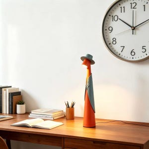 May include: A whimsical orange desk lamp with a gray hat and vest design. The lamp is on a wooden desk with books, a clock, and a small plant. The lamp's design is unique and adds a touch of personality to any workspace.