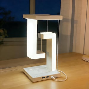 May include: A white, modern desk lamp with a unique, floating design. The lamp has a rectangular base with USB ports and a wireless charging pad. The light is suspended by thin wires, creating a minimalist aesthetic. The lamp is plugged in with a white cable.