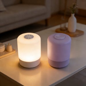 Puede incluir: Dos lámparas de mesa cilíndricas. Una está encendida, emitiendo un suave brillo blanco, y la otra es de color lavanda apagado. Ambas lámparas tienen un panel de control circular en la parte superior y una base blanca. Las lámparas están sobre una mesa blanca.