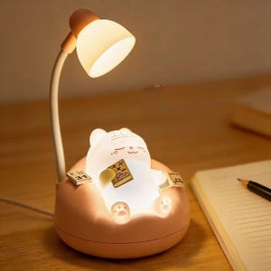 Puede incluir: Una lámpara de escritorio caprichosa con un oso de dibujos animados leyendo un libro, iluminada por una luz suave y cálida. La lámpara tiene una base de color melocotón y un cuello blanco flexible, coronado por una pantalla de color melocotón. El oso sostiene un libro y tiene una expresión serena.