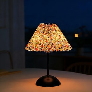 May include: A small table lamp with a black base and stem, topped with a floral patterned lampshade. The lampshade is a mix of orange, red, blue, and yellow flowers. The lamp is on a white surface with a dark background.