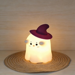 Puede incluir: Una lámpara de noche con forma de fantasma sonriente, con un sombrero de bruja morado. El fantasma tiene ojos negros y mejillas rosadas. Se encuentra sobre una estera redonda tejida. La luz emite un brillo suave y cálido.