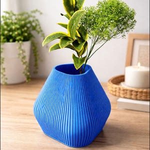 以下が含まれることがあります： テクスチャ加工されたリブデザインの鮮やかな青い花瓶。花瓶には、緑と黄色の縁取りの葉と、緑の葉の小枝が入っています。花瓶は木製の表面にあり、背景には他の植物や装飾があります。