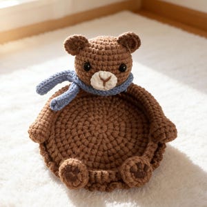 May include: A handmade brown crochet bear-shaped basket with a light blue scarf. The bear has small black eyes, a beige muzzle, and small brown ears. The basket is sitting on a white, fluffy rug.