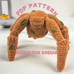 Puede incluir: Una criatura tejida a ganchillo, de color marrón, con forma de araña y detalles verdes. El texto "PDF PATTERN" y "ROCKY THE ERIDIAN" se muestran en rosa. El juguete tiene múltiples patas y un cuerpo redondeado.