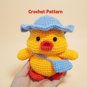 Op de afbeelding: Een handgemaakte gele gehaakte eend met een blauwe hoed en een blauwe tas. De eend heeft oranje voeten en een rode snavel. De tekst "Crochet Pattern" staat in het rood bovenaan de afbeelding.
