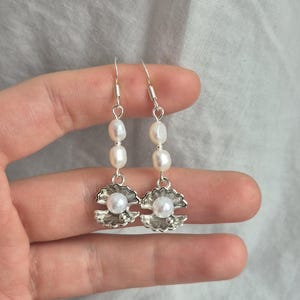 May include: A pair of silver-toned drop earrings featuring two small white oval pearls stacked vertically above a silver-toned metal charm shaped like an open clam shell, which contains a single round white pearl at its centre.