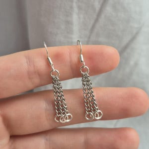 May include: A pair of silver-toned dangle earrings featuring three delicate, thin metal chains suspended from a standard hook wire. The chains hang vertically and are connected at the bottom by small, circular jump rings, creating a layered effect.