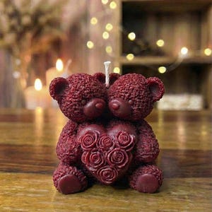 May include: A burgundy candle in the shape of two teddy bears cuddling, holding a heart decorated with roses. The candle has a wick and is placed on a wooden surface. The background is blurred with warm lighting.
